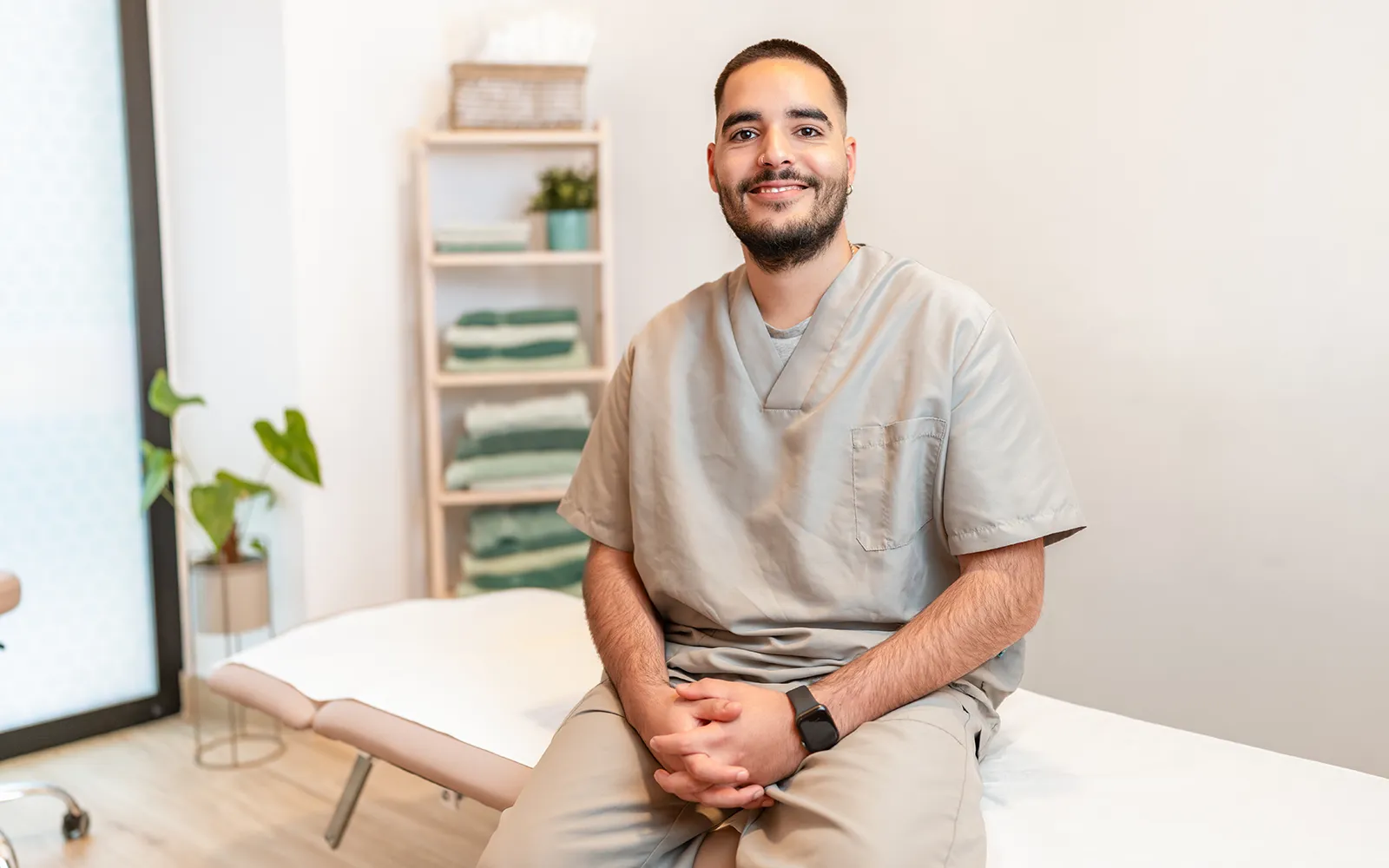 A massage therapist sits on the edge of a massage table, smiling for a photo.