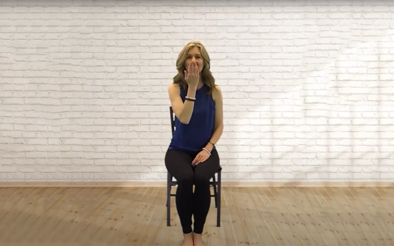 MA21_ChairYogaUjjayBreath_Main A woman sits on a chair with her legs relaxed, one hand on her lap and one in front of her mouth as she inhales.