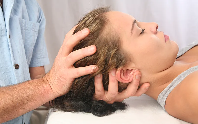 A massage therapist uses his hands on a supine female client's neck and head.